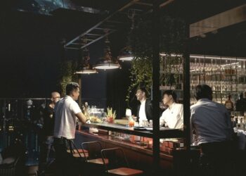 A man enjoying a craft cocktail at one of the best rooftop bars in NYC with the Manhattan skyline glowing behind her at dusk