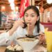 woman-eating-soup-dumplings-unsplash - NYCwebzine Una mujer levanta cuidadosamente una sopa con fideos con palillos en una mesa en Chinatown de Flushing, Nueva York