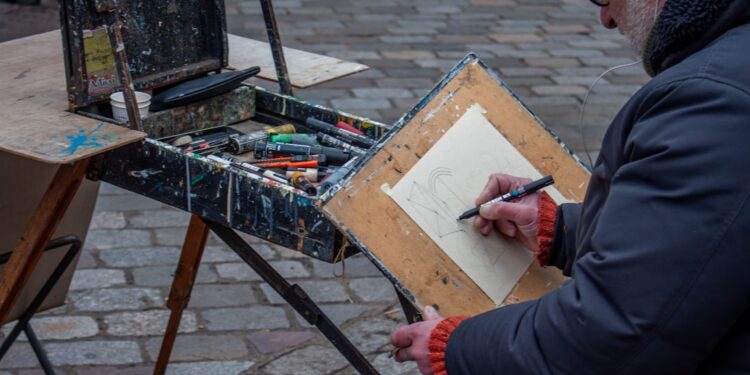 A man sits outdoors with a sketchbook, taking part in free art classes in a New York City park setting