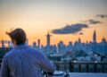 man-rooftop-skyline-cocktail-unsplash - NYCwebzine A man enjoying a cocktail on a Manhattan rooftop terrace at the city's top private clubs with a skyline view behind him