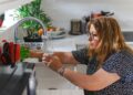 woman-organizing-glass-jars-kitchen-unsplash - NYCwebzine A woman arranging labeled glass jars on an open shelf as part of building a lower-waste kitchen in a New York City apartment